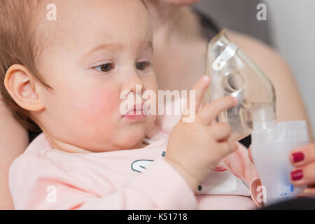 Un anno di età Baby girl tenendo l'inalatore, sua madre mentre tiene il suo nei bracci e consolante il bambino malato Foto Stock