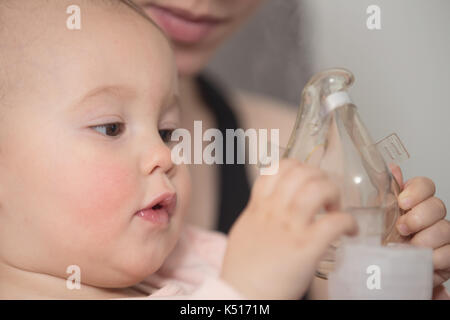 Un anno di età Baby girl tenendo l'inalatore, sua madre mentre tiene il suo nei bracci e consolante il bambino malato Foto Stock