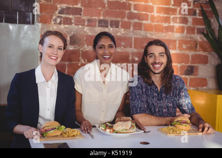 Ritratto di sorridere giovani amici seduti con il cibo in tavola contro un muro di mattoni in coffee shop Foto Stock