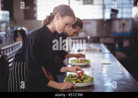 Vista laterale di giovani camerieri preparare insalata fresca piastre mentre è in piedi in cucina commerciale presso la caffetteria Foto Stock