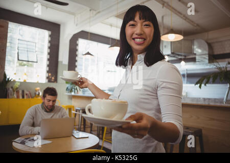 Ritratto di sorridente cameriera holding tazze da caffè con uomo utilizzando laptop a tavola in cafe Foto Stock