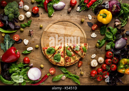 Lay piatto con la pizza italiana le fette su tavola di legno con vari ortaggi freschi e le erbe sul tavolo Foto Stock