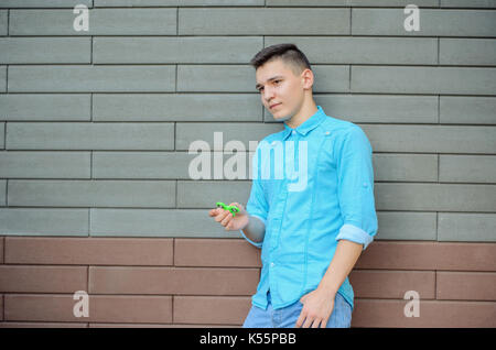 Pensieroso giovane uomo sull'azzurro camicia e jeans blu mano azienda gadget popolari agitano: spinner. adolescente giocando con il filatore verde all'aperto su Foto Stock