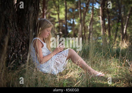 Lunghi capelli biondi carina ragazza in abito bianco seduto appoggiato la schiena contro tree guardando pigna nelle sue mani. Foto Stock