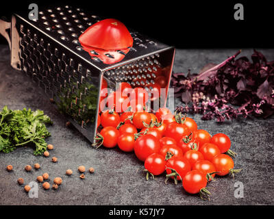 Preparazione della ricetta succo di pomodoro. pomodoro grande e vecchia grattugia fino a piccoli uva pomodori ciliegia sul retro vintage rustico in pietra grigia sullo sfondo. Foto Stock