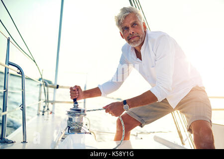 Uomo maturo in piedi sul ponte della sua barca utilizzando un argano mentre sono fuori per una vela di un pomeriggio soleggiato Foto Stock