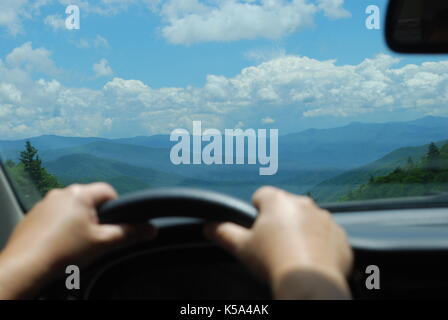 Fantastica vista attraverso l'auto finestra anteriore sulle montagne con cielo blu e nuvole bianche. mani del conducente sul volante Foto Stock