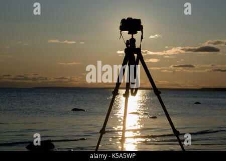 Fotocamera con treppiede oltre il tramonto vicino acqua di mare Foto Stock