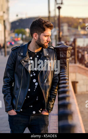 Un bel giovane hipster uomo appoggiato alla recinzione e guardando intorno al tramonto Foto Stock