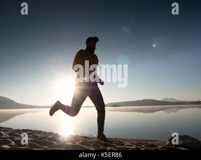 Uomo che corre sulla spiaggia contro lo sfondo di un bel tramonto. sabbia del lago di montagna con piede profondo sentieri. Foto Stock