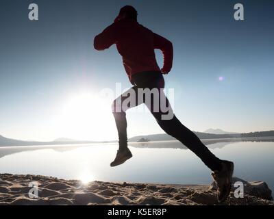 Uomo che corre sulla spiaggia contro lo sfondo di un bel tramonto. sabbia del lago di montagna con piede profondo sentieri. Foto Stock