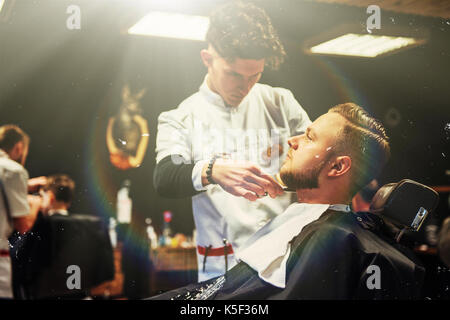 Il barbiere uomo nel processo di taglio della barba di client tosatrici elettriche in barberia. Foto Stock