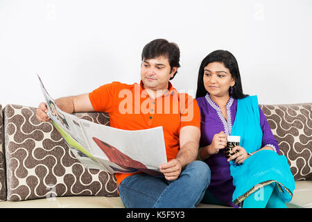 Indian giovane sul divano a bere caffè e quotidiano di lettura a casa Foto Stock