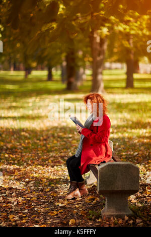 Curly redhead donna leggere il libro nel parco in autunno Foto Stock