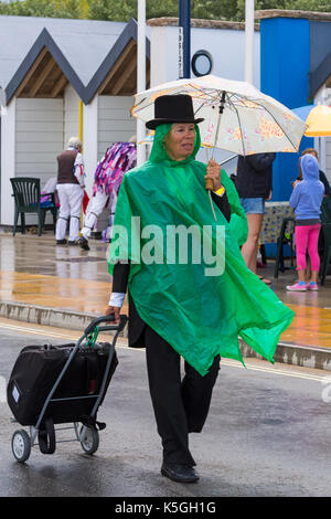 Swanage, Dorset, Regno Unito. 9 Sep, 2017. Regno Unito meteo: heavy rain e tuoni a Swanage. Tirando la donna valigia trolley e indossare top hat e poncho verde azienda ombrello passeggiate lungo il lungomare spiaggia del passato di capanne. Credito: Carolyn Jenkins/Alamy Live News Foto Stock