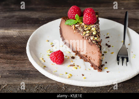 Fetta di Cheesecake al cioccolato decorate con pistacchi, lamponi e foglia di menta sulla piastra bianca su sfondo di legno. Foto Stock