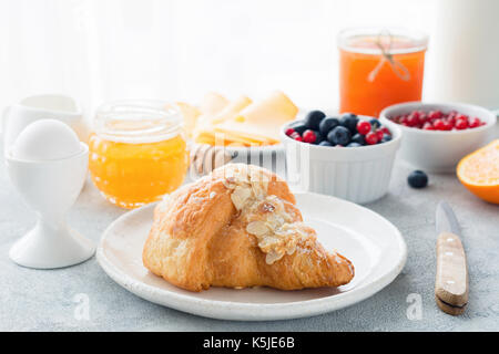 Croissant alle mandorle, miele di mirtilli freschi, arancio, marmellata, uve secche di Corinto, uovo sodo e una bottiglia di latte sulla tavola . closeup vista della colazione continentale. cop Foto Stock