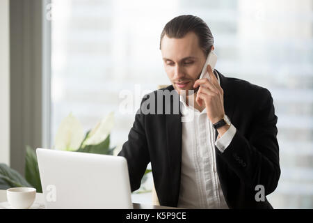 Giovane imprenditore in ufficio guardando laptop effettuare chiamate telefoniche. Foto Stock