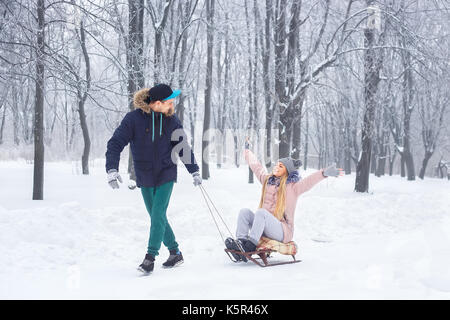 Felice inverno giovane uomo slittino su sled donna per le vacanze invernali in boschi innevati. Divertimento giovane giocoso insieme all'aperto. persone, stagione invernale, leisur Foto Stock