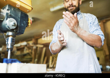 Basso angolo di vista fiducioso artigiano di segatura di scuotimento da mani dopo il completamento dei lavori con il trapano pressa, sfondo sfocato Foto Stock