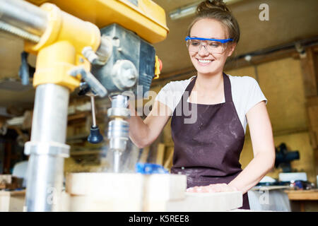 Attraente giovane falegname con sorriso toothy con trapano pressa per eseguire fori in asse in legno, sfondo sfocato Foto Stock