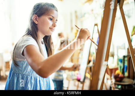 La vista del profilo della graziosa bambina indossa abiti a strisce che realizza le sue idee sulla tela con l aiuto del pennello e acquerelli, interni di art studio Foto Stock