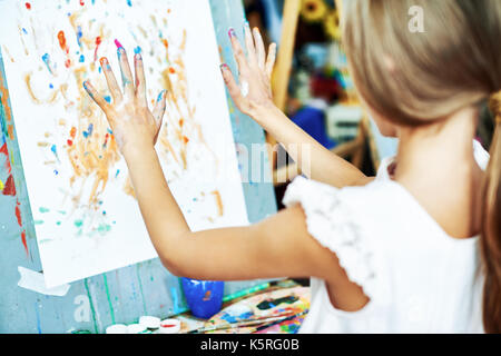 Vista posteriore di un irriconoscibile bambina permanente al cavalletto e facendo finger-pittura, le mani sono ricoperte con acquarelli Foto Stock