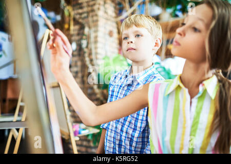 Vista di profilo di bella ragazza seduta di fronte a cavalletto e disegno mentre il suo compagno di classe in piedi accanto a lei e guardando il dipinto incompiuto con int Foto Stock