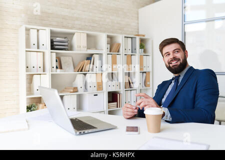 Ritratto di successo imprenditore barbuto sorridente felicemente mentre si lavora in un ufficio moderno Foto Stock