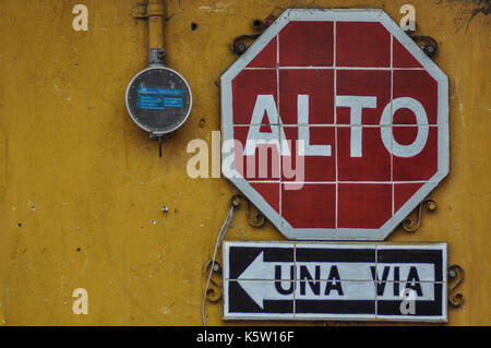 Un segnale di stop rosso e bianco con la scritta "alto" è montato su una parete gialla sopra un cartello bianco e nero che indica "una via", che si traduce in "a senso unico" Foto Stock