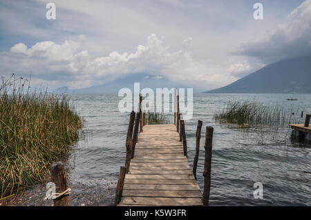 Un molo di legno si estende in un lago calmo circondato da montagne e sotto un cielo nuvoloso. Foto Stock