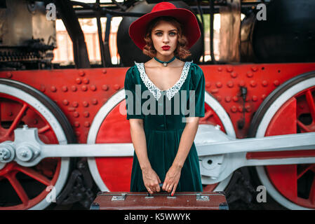 Woman in Red Hat con la valigia in mano contro vintage locomotiva a vapore, ruote rosse closeup. Il vecchio treno. Motore ferroviaria, viaggio ferroviario Foto Stock