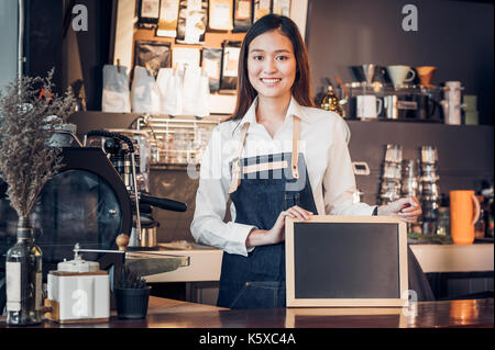 Femmina asiatica barista indossare jean grembiule e azienda lavagna vuota menu al banco bar con sorriso faccia,servizio di caffetteria concetto,proprietario business start up,moc Foto Stock