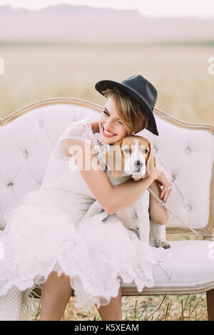 Il Sorridente sposa cercando altrove è abbracciando il cagnolino seduto sul bianco divano vintage presso lo sfondo del campo. close-up verticale. Foto Stock