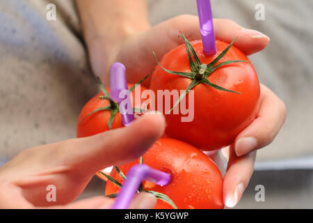 Tre mature pomodori rossi con cannucce nelle mani della donna su sfondo chiaro. succo di frutta fresco cibo sano concetto. il pomodoro in un recipiente con del succo di frutta fresco Foto Stock