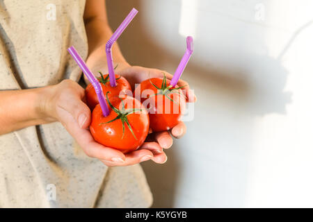 Tre mature pomodori rossi con cannucce nelle mani della donna su sfondo chiaro. succo di frutta fresco cibo sano concetto. il pomodoro in un recipiente con del succo di frutta fresco Foto Stock