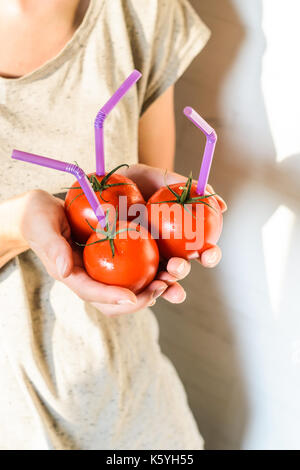 Tre mature pomodori rossi con cannucce nelle mani della donna su sfondo chiaro. succo di frutta fresco cibo sano concetto. il pomodoro in un recipiente con del succo di frutta fresco Foto Stock