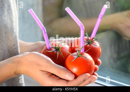 Tre mature pomodori rossi con cannucce nelle mani della donna su sfondo chiaro. succo di frutta fresco cibo sano concetto. il pomodoro in un recipiente con del succo di frutta fresco Foto Stock