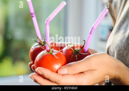 Tre mature pomodori rossi con cannucce nelle mani della donna su sfondo chiaro. succo di frutta fresco cibo sano concetto. il pomodoro in un recipiente con del succo di frutta fresco Foto Stock