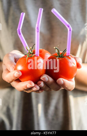 Tre mature pomodori rossi con cannucce nelle mani della donna su sfondo chiaro. succo di frutta fresco cibo sano concetto. il pomodoro in un recipiente con del succo di frutta fresco Foto Stock