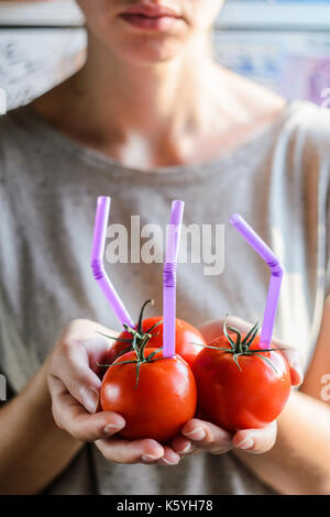 Tre mature pomodori rossi con cannucce nelle mani della donna su sfondo chiaro. succo di frutta fresco cibo sano concetto. il pomodoro in un recipiente con del succo di frutta fresco Foto Stock