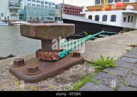 Rusty bollard con verde linea di ormeggio che conduce a una nave lungo il molo e moderni edifici in background, Bremerhaven porto di pesca, Germania Foto Stock