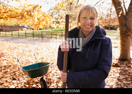 Ritratto di donna matura a rastrellare foglie di autunno in giardino Foto Stock