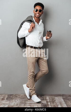 A piena lunghezza Ritratto di un sorridente uomo africana in occhiali da sole azienda tazza di caffè per andare isolate su uno sfondo grigio Foto Stock