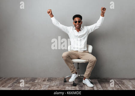 Felice eccitato uomo africana in occhiali da sole per celebrare il successo mentre è seduto in una sedia isolate su uno sfondo grigio Foto Stock