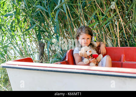 Poco carino bambina si siede in una barca sul lago. Ha conseguito il suo giocattolo preferito in mano. l'infanzia, crescendo, giocando. copia incolla Foto Stock