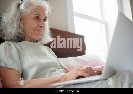 Donna anziana che stabilisce nel letto tramite computer portatile Foto Stock