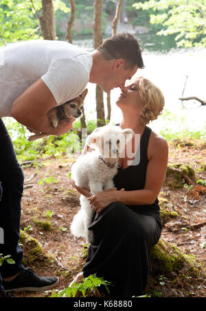 Un uomo e una donna toccare nasi affettuosamente nel bosco, tenendo i loro cani, cane bianco che guarda fuori Foto Stock