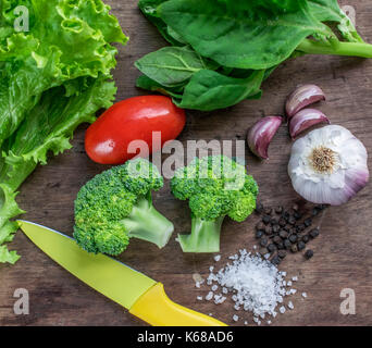 Verdure verdi e spezie, mostrando le lattughe e spinaci, viola aglio, due teste di broccoli, un rosso pomodoro, il pepe nero e il sale, sullo sfondo di legno Foto Stock