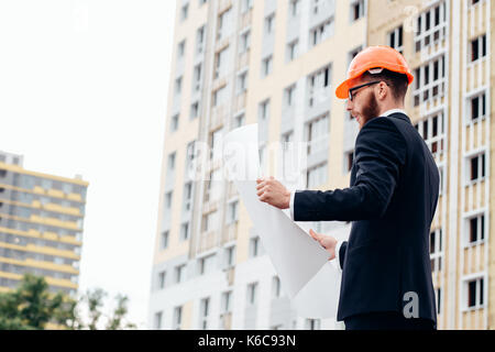 Ritratto di un architetto builder studiando piano di disposizione delle camere, serio ingegnere civile che lavora con i documenti sul sito in costruzione. Foto Stock